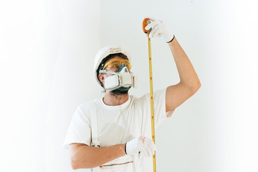pexels-photo-4981777 A construction worker wearing safety equipment measures with a tape against a light background.